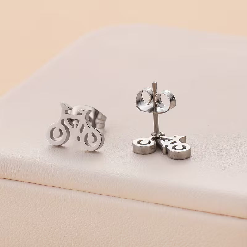 A pair of silver bicycle stud earrings shown on a light beige textured surface, with one earring facing forward and the other showing the post and butterfly back.