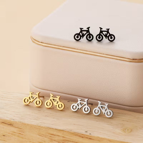 Bicycle-shaped earrings in black, gold, and silver on a beige cushion with wooden surface.